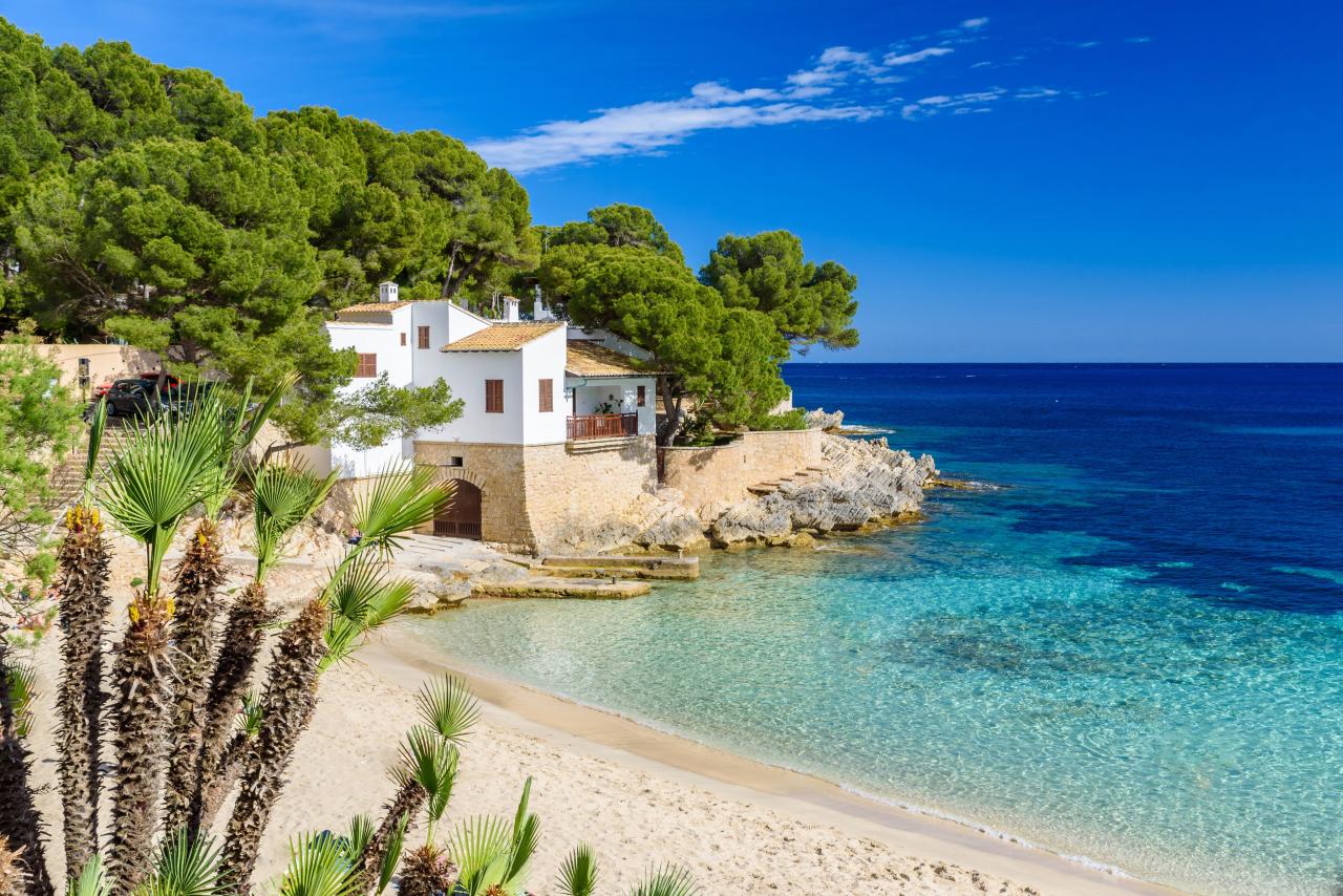 Sonniger Strand der Cala Gat in Cala Ratjada auf Mallorca mit Wohnhaus im Hintergrund.
