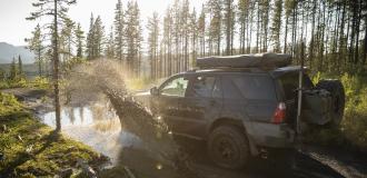 Ein Geländewagen fährt durch eine Pfütze auf einem Waldweg mit Bergen und Bäumen im Hintergrund.