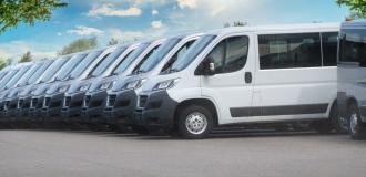 Eine Reihe weißer Kleinbusse steht auf einem Parkplatz bei sonnigem Wetter und leicht bewölktem Himmel.