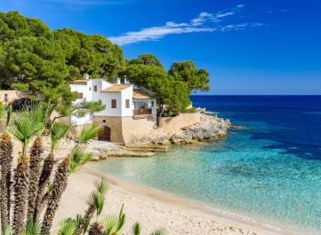 Sonniger Strand der Cala Gat in Cala Ratjada auf Mallorca mit Wohnhaus im Hintergrund.