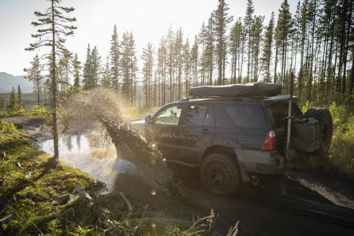 Ein Geländewagen fährt durch eine Pfütze auf einem Waldweg mit Bergen und Bäumen im Hintergrund.