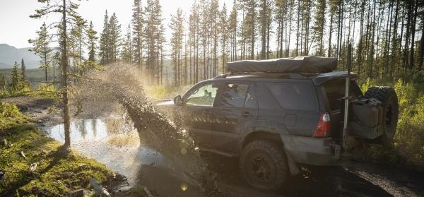 Ein Geländewagen fährt durch eine Pfütze auf einem Waldweg mit Bergen und Bäumen im Hintergrund.
