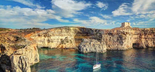 Comino Tower auf Malta vom Meer gesehen, mit Klippen und Segelboot im Vordergrund.