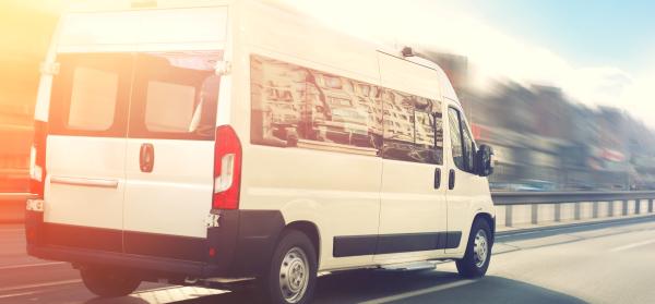 Ein weißer Kleinbus fährt schnell auf einer Straße in der Stadt, mit Gebäuden im Hintergrund.