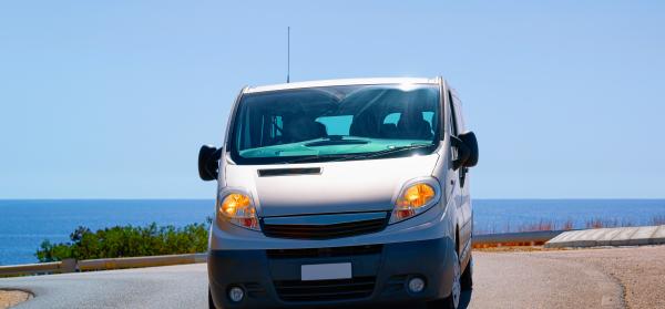 Ein weißer Van fährt auf einer kurvigen Küstenstraße mit Blick auf das Meer und den blauen Himmel im Hintergrund.