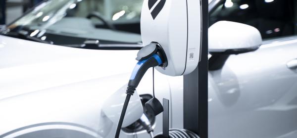 Eine Ladestation für E-Autos mit angeschlossenem Ladekabel, im Hintergrund ein weißes Elektroauto.