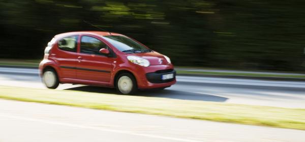 Ein roter Kleinwagen fährt schnell auf einer Straße mit einer grünen Landschaft im Hintergrund.