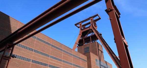 Backsteingebäude einer ehemaligen Zeche mit rostbraunem Förderturm und Rohrkonstruktion in Essen vor blauem Himmel.