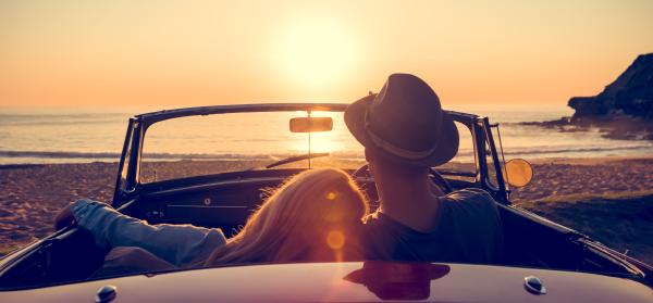Ein Paar sitzt in einem Cabrio am Strand und schaut gemeinsam auf den Sonnenuntergang über dem Meer.