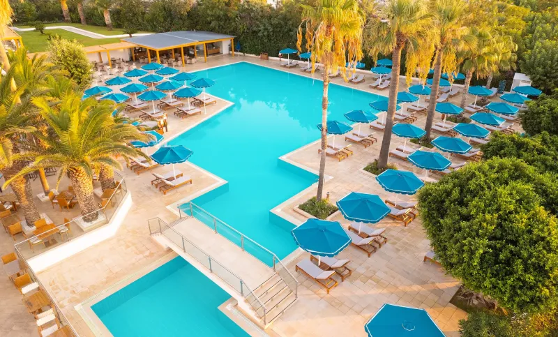 Großzügiger Poolbereich im Mitsis Faliraki Beach Hotel auf Rhodos mit Liegen, Palmen und blauen Sonnenschirmen bei Sonnenaufgang