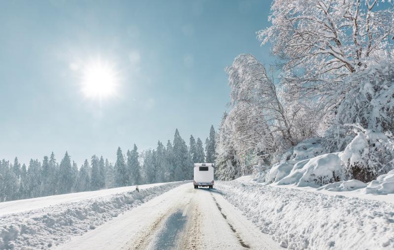 weißes Wohnmbil fährt auf zugeschneiter Straße durch eine verschneite Winterlandschaft
