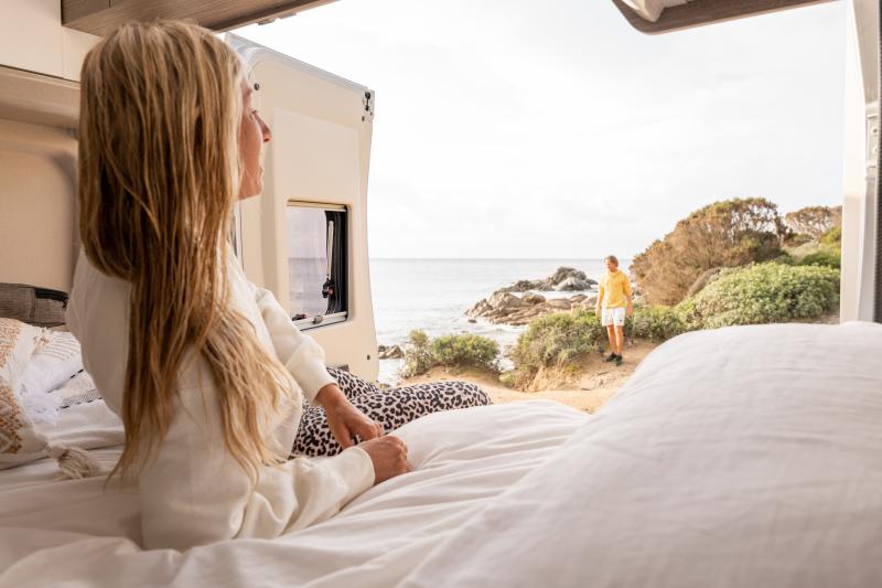 Eine Frau liegt in ihrem Wohnmobil und lächelt durch die geöffneten Hecktüren einem Mann am Meer zu.