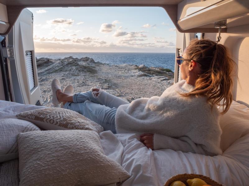 Eine im Camper liegende Frau schaut durch die geöffneten Hecktüren nach draußen auf das Meer, während die Sonne untergeht.