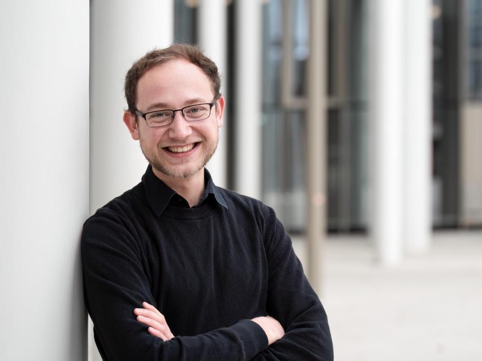 Simon Pietras lächelt vor moderner Architektur mit schwarzem Pullover und Brille.